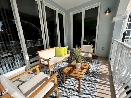 A cozy balcony with wooden furniture, cushioned seating, a patterned rug, and a small table with potted plants. Sunlight streams in.