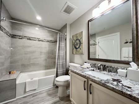 A modern bathroom features a bathtub, shower, toilet, and a granite countertop vanity with a large mirror and nautical decor.