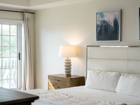 Bright bedroom with an open window, white bed, bedside lamp, dresser, and framed ocean-style artwork on the wall.