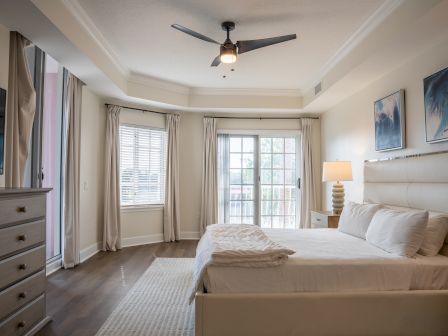 A bright modern bedroom with a ceiling fan light, large windows with curtains, framed wall art, and a neatly made bed.