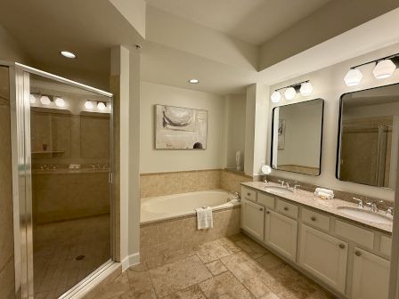 A bathroom featuring a glass shower, bathtub, dual sinks with mirrors, beige tiles, and overhead lights.