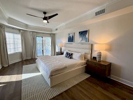 A modern bedroom with a bed, two nightstands, lamps, artwork, and a ceiling fan. Large windows let in natural light, with curtains.