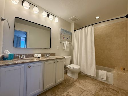 A clean bathroom features a large mirror, double sink, tissue box, towels, toilet, and a shower with a white curtain.