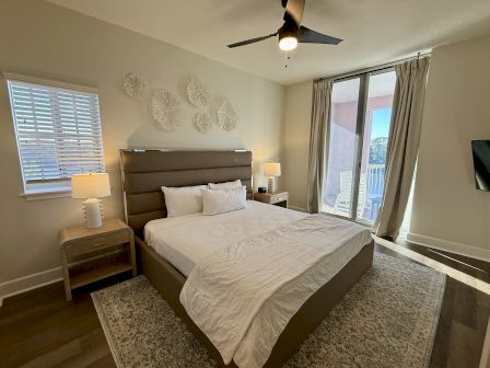 A modern bedroom with a large bed, lamps on side tables, decorative wall art, a ceiling fan, and window blinds on a sunny day.