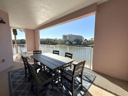 A balcony features a dining table with chairs, lounger, overlooking a serene water view with a building in the background under clear skies.