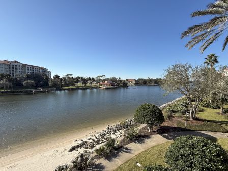 A scenic view of a waterfront with a sandy beach, lush greenery, trees, and a building in the background under a clear blue sky.
