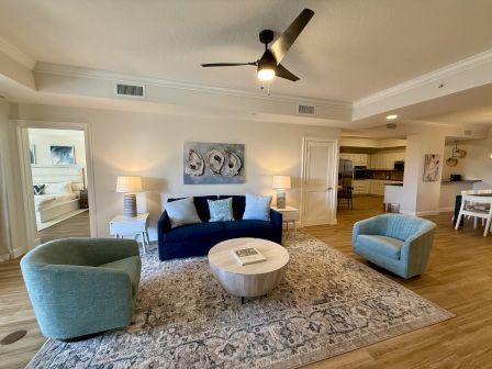 A cozy living room with a blue sofa, round table, two blue chairs, artwork, lamps, and visible kitchen and bedroom areas.