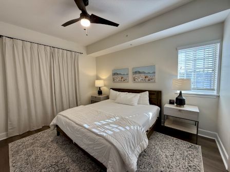 A bedroom with a bed, two side tables with lamps, a ceiling fan, artwork on the wall, and a window with blinds.