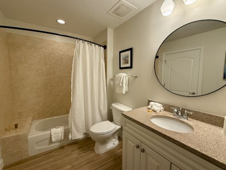 A clean bathroom features a bathtub with a curtain, a toilet, a countertop with a round mirror, towels, and light fixtures.