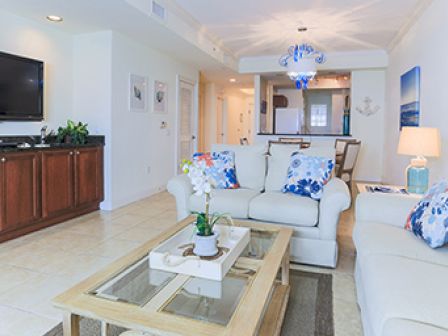 A cozy living room with a TV, white sofas, blue-themed decor, a glass coffee table, a lamp, and an open kitchen in the background.