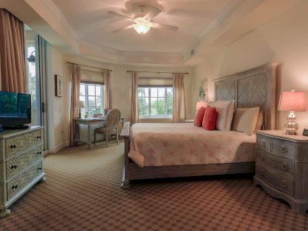 A cozy bedroom with a double bed, dresser, TV, desk by windows, and ceiling fan. Soft lighting and neutral tones create a calm ambiance.