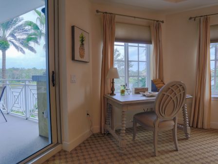 A cozy room with a wooden desk, chair, and lamp, overlooking a balcony and palm trees through the window and glass door.