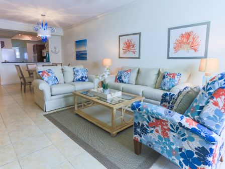 A living room with beige sofas, colorful cushions, a floral armchair, a glass coffee table, wall art, and a blue chandelier.