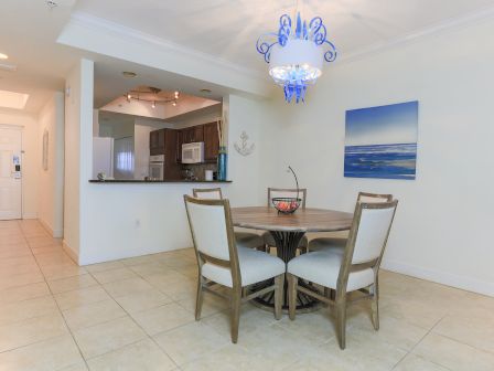 A dining area with a round table and four chairs, near a kitchen with wooden cabinets, under a blue chandelier.