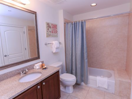 The image shows a bathroom with a sink, mirror, toilet, and bathtub with a tiled wall. A "Sand & Sea" sign decorates the wall.