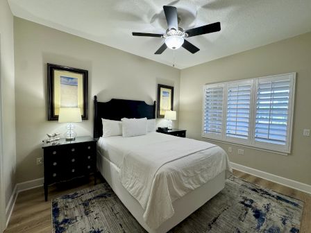 A bedroom with a bed, two nightstands, framed art, lamps, a ceiling fan, windows with shutters, and a patterned rug on a wooden floor.