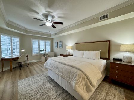 A bedroom with a large bed, two nightstands, lamps, a desk with a chair, and a ceiling fan in a well-lit room with wooden floors.