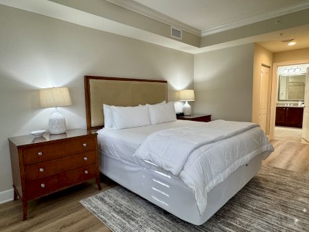 A neatly made bed with white linens is flanked by wooden nightstands and lamps; a bathroom is visible in the background.