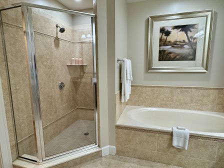 A bathroom with a glass-enclosed shower, bathtub, towels, and wall art with a nature scene. The decor features beige tiles.