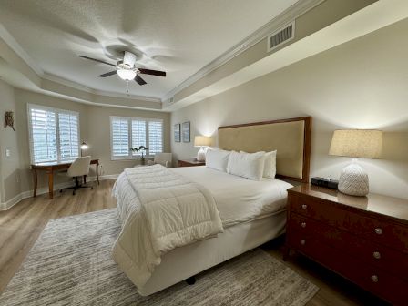 A cozy bedroom with a large bed, two nightstands, lamps, a ceiling fan, a desk, chair, and windows with blinds.
