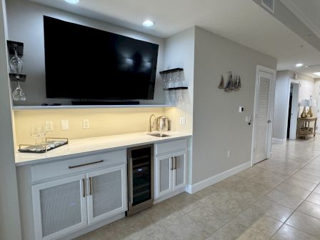 A home bar area with a large TV, shelves with glasses, sink, wine fridge, and decorations, set in a modern interior space.