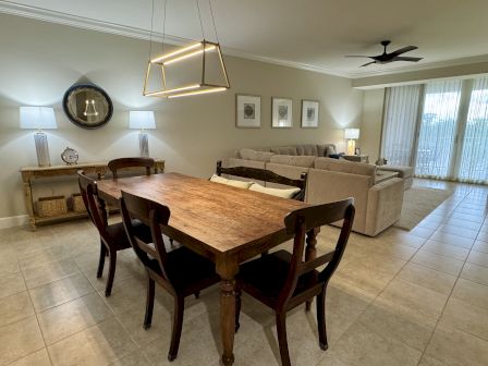 A cozy, modern living and dining area with a wooden table, ambient lamps, beige sofas, and large windows with sheer curtains.