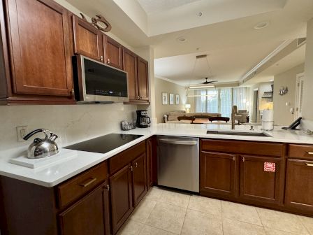 A modern kitchen with wooden cabinets, stove, kettle, coffee maker, and dishwasher. The living area is visible in the background.