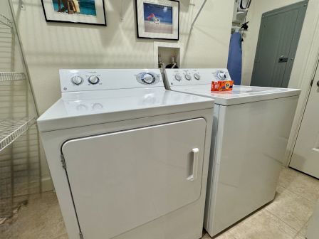 A laundry room features a washer, dryer, shelf, ironing board, framed photos, and a Tide detergent box on a tile floor.