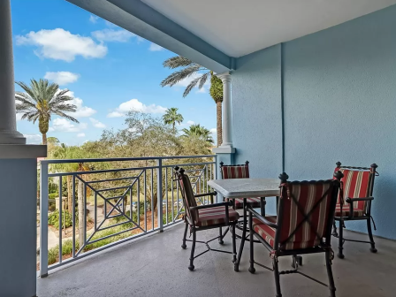 A balcony with a table and four chairs, overlooking a garden with palm trees and a clear blue sky in the background.