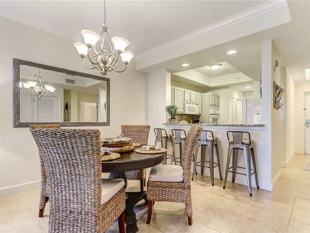 A dining area with wicker chairs and a round table, a kitchen with bar stools, and a large mirror reflecting a chandelier can be seen.