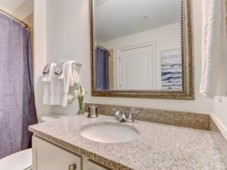 A bathroom with a granite countertop, sink, mirror, towel rack, and a blue shower curtain. A small vase with flowers is also present.