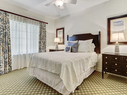 A cozy bedroom with a large bed, decorative pillows, elegant nightstands, lamps, framed art, and patterned curtains by the window.