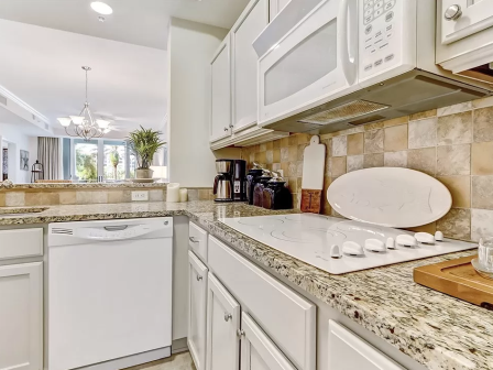 A bright, modern kitchen with granite countertops, white cabinets, a stovetop, microwave, dishwasher, coffee maker, and decor ending the sentence.