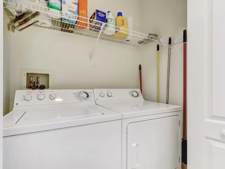 A laundry area with a washer, dryer, cleaning supplies on a shelf, and brooms against the wall.