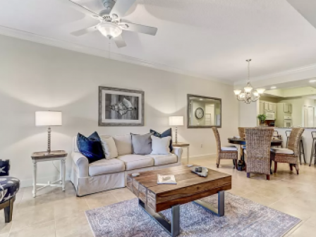 A modern living room with a sofa, coffee table, and dining area; neutral tones and ceiling fan complete the decor.
