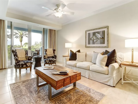 A cozy living room with a sofa, patterned chairs, coffee table, lamps, and art. Large windows reveal a view of trees and a balcony.