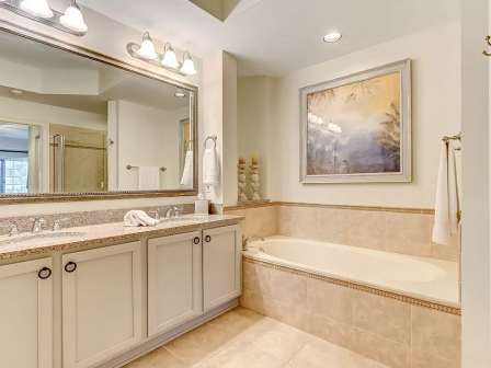 The image shows a bathroom with a large mirror, twin sinks, a bathtub, a painting, and warm lighting for a cozy ambiance.