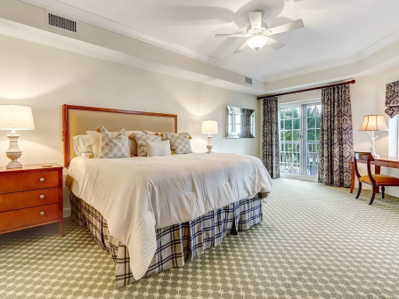 A cozy bedroom with a large bed, side tables, lamps, a desk, and curtains, featuring a ceiling fan and a view through French doors.