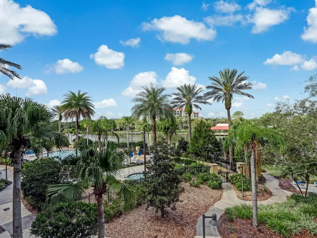 The image shows a tropical setting with palm trees, pathways, manicured gardens, and a pool under a bright blue sky with fluffy clouds.