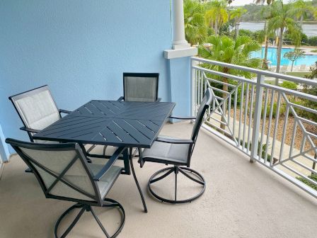 A balcony with a black patio table and four chairs, overlooking a pool surrounded by palm trees and greenery.