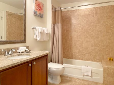 A bathroom with a granite countertop, sink, mirror, toilet, towels, artwork, and a bathtub with a tiled shower area.