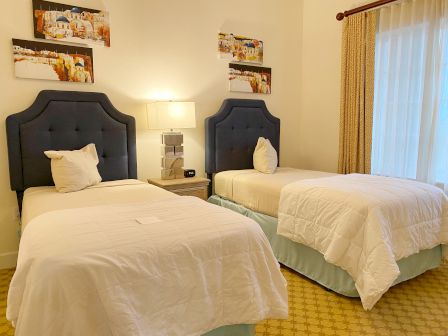 The image shows a hotel room with two single beds, a bedside table, a lamp, and artwork on the wall.