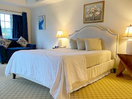 A cozy bedroom featuring a bed with white linens, lamps on side tables, artwork on the walls, and a sitting area by the window.