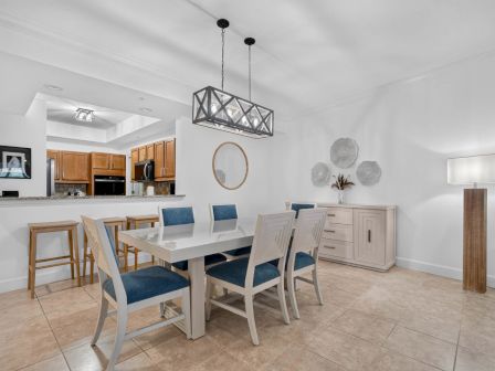 A bright modern dining room with a white table, blue chairs, and a view into a kitchen with wooden cabinets.