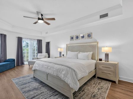 A bright modern bedroom with a large bed, nightstands, lamps, ceiling fan, and floor-to-ceiling curtains by a sunny window.