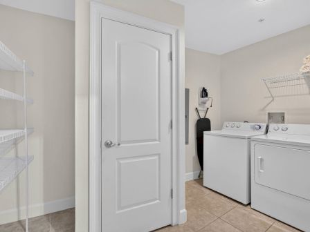 A bright laundry room with a white washer and dryer, a storage wire shelf, and an ironing board beside a utility closet door.