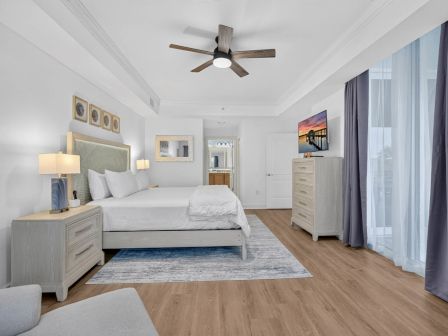 A modern bright bedroom with a large bed, ceiling fan, dressers, wall art, and blue curtains by the window.