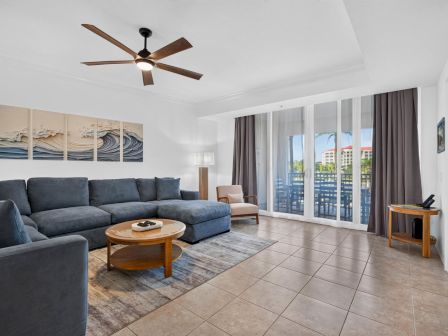 Bright modern living room with blue sectional sofa, coffee table, ceiling fan, and large sliding doors to a balcony view.