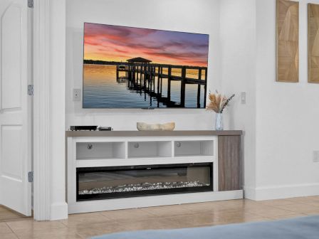 A bright modern living room shows an electric fireplace, a sunset pier wall photo, and simple decor on a white cabinet, .