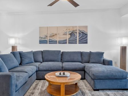 Bright living room with a blue sectional sofa, round wooden coffee table, wall ocean artwork, lamps, and a ceiling fan.